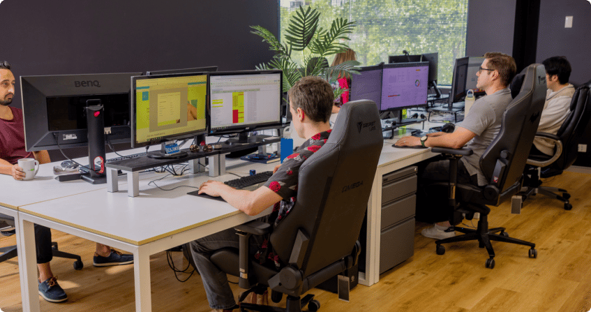 four team members working at dual-monitor workstations in an open-plan office with timber floors and natural light, reviewing campaign data and audit spreadsheets.