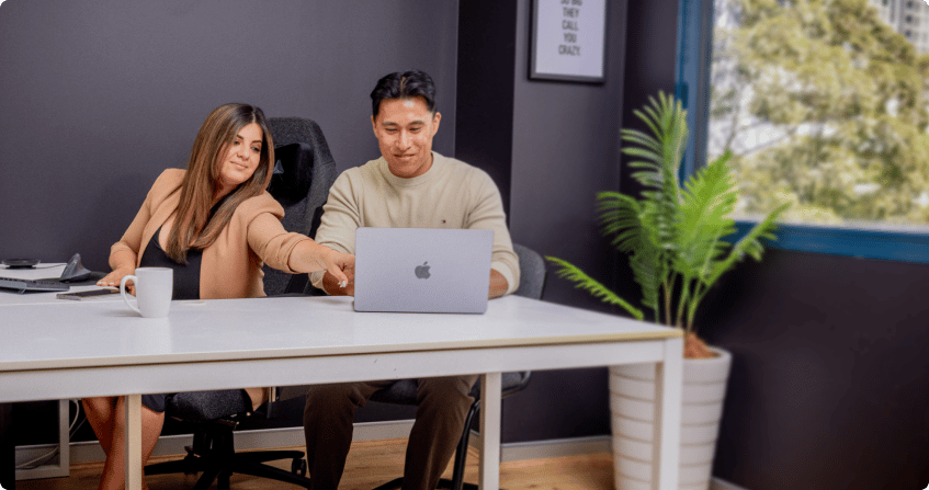 two team members sitting at a white desk reviewing campaign data on a laptop, one pointing at the screen, in a modern office with dark walls, a framed quote, and a potted palm near the window.