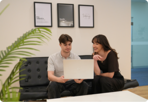 Two Click Click Media team members review work on a laptop together on a black leather couch in the office.