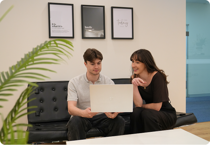 Two Click Click Media team members review work on a laptop together on a black leather couch in the office.