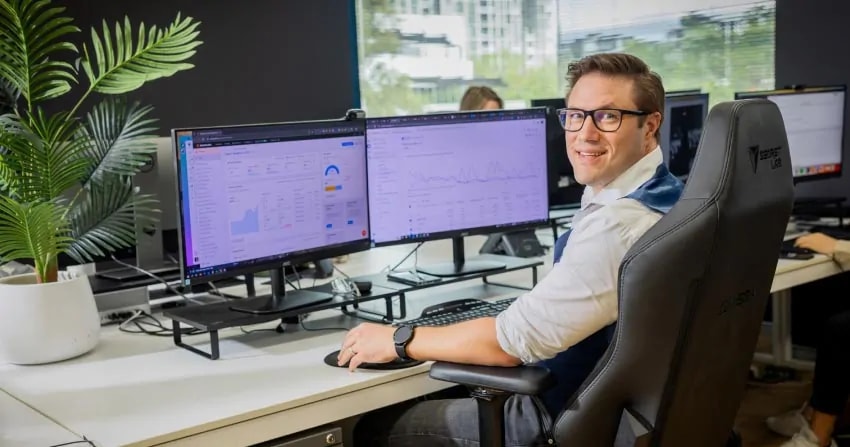 team member sitting at a dual-monitor workstation with semrush analytics dashboards open on screen, wearing glasses and a white shirt, with a potted plant and natural light from the office windows.
