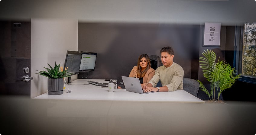two google ads specialists sitting side by side at a white desk reviewing campaign data on a laptop in a modern office with dark walls and natural light.